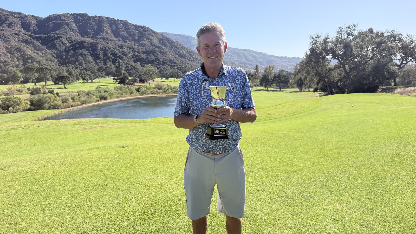 John Pate holding the Ojai Senior Cup Super Senior trophy on the tee at Soule Park, with a pond and mountains behind him